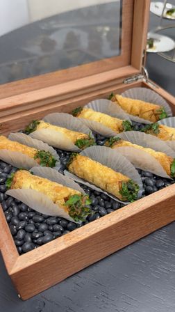 Crispy bite-sized fried rolls garnished with chopped herbs, arranged in paper liners on a bed of black beans inside a wooden display box — gourmet catered hors d'oeuvres.