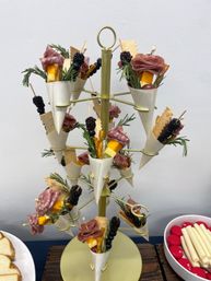 Tiered gold stand of mini charcuterie cones — salami roses, cheddar cubes, crackers, pretzels, blackberries and rosemary sprigs for a party appetizer display.