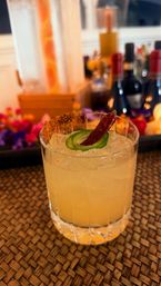 Spicy jalapeño‑chili craft cocktail in a rocks glass with chili‑rimmed salt and crushed ice on a woven placemat, blurred bar bottles and floral garnish behind