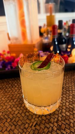 Spicy jalapeño‑chili craft cocktail in a rocks glass with chili‑rimmed salt and crushed ice on a woven placemat, blurred bar bottles and floral garnish behind