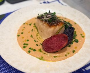 Seared white fish fillet topped with microgreens, plated on a creamy pink sauce with green herb oil droplets and roasted purple-and-red potatoes on a white plate