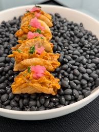 Row of bite-sized crispy chicken tacos topped with pickled pink onions and cilantro, presented on a bed of black beans in a white bowl — Mexican-style appetizer