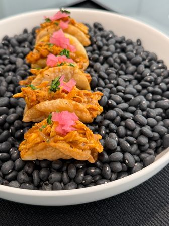 Row of bite-sized crispy chicken tacos topped with pickled pink onions and cilantro, presented on a bed of black beans in a white bowl — Mexican-style appetizer