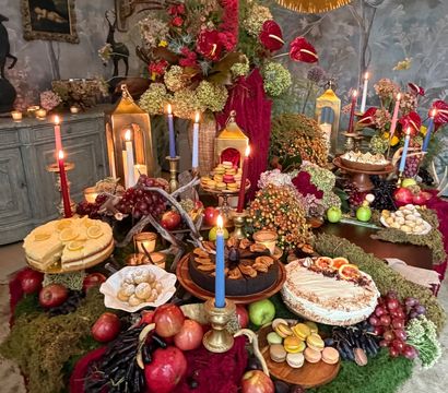 Opulent autumn tablescape in a moody vintage room: moss-draped table topped with lit tapers and lanterns, lush floral centerpiece, cakes, macarons, apples, grapes and assorted pastries.