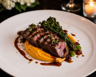 Sliced grilled steak over creamy sweet potato purée with charred broccolini, microgreens and glossy balsamic reduction on a white plate — elegant candlelit restaurant entrée.