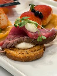Gourmet appetizer: thin rare roast beef on multigrain crostini with a dollop of creamy cheese and microgreens, plated beside two red-coated croquettes topped with blueberries.