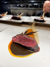 Close-up of a medium-rare seared steak slice on a dark reduction and bright orange purée, garnished with purple microgreens and a corn-husk side on a white plate at an open-kitchen fine-dining pass.