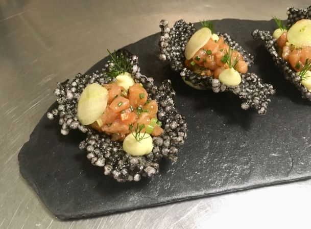 Salmon tartare amuse-bouches on black puffed rice crisps, topped with thin radish slices, fresh dill and lemon aioli dollops on a slate board