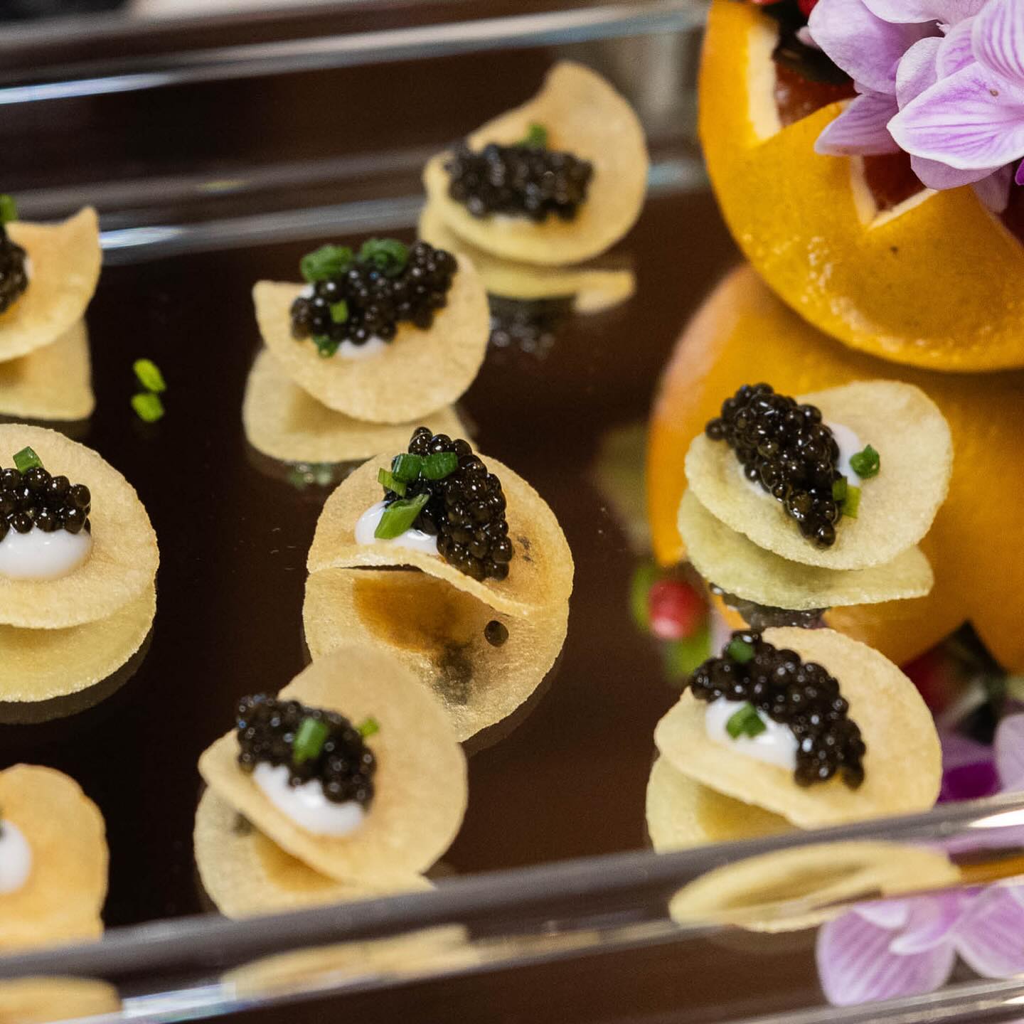 Tiny gourmet caviar canapés — glossy black caviar on crème fraîche-topped potato crisps, sprinkled with chives on a mirrored serving tray with a floral garnish