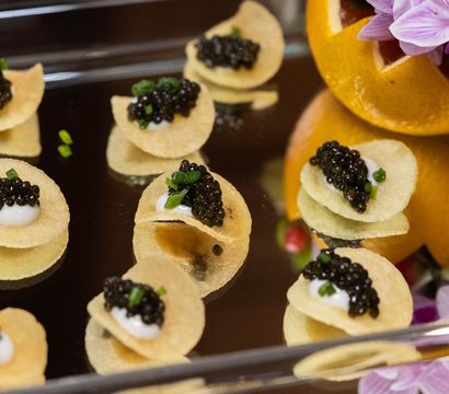 Tiny gourmet caviar canapés — glossy black caviar on crème fraîche-topped potato crisps, sprinkled with chives on a mirrored serving tray with a floral garnish