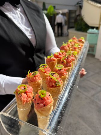 Server in black vest holding a clear tray of bite-sized tuna tartare cones garnished with green onion curls and black sesame at an outdoor catered event