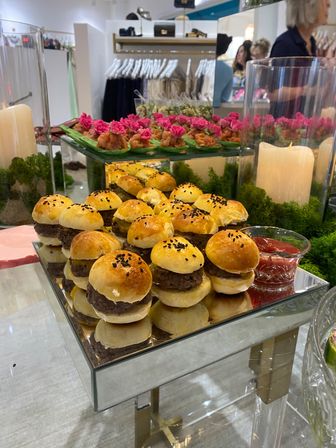 Mirror tray of bite-sized beef sliders on sesame-topped brioche buns with ketchup, displayed beside pink-flowered canapés and candles, set against clothing racks in a boutique event setting.