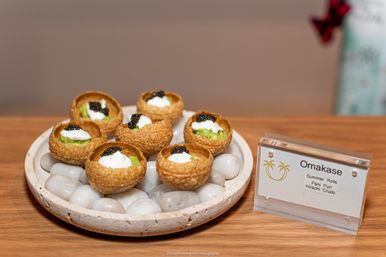 Seven bite-sized pani puri shells filled with avocado, cream and black caviar on a stone-lined ceramic plate beside an 'Omakase' menu card on a wooden table.