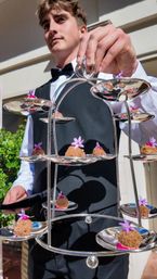 Waiter in black vest and bow tie holding a tiered silver serving stand with bite-sized crunchy canapés topped with small purple edible flowers on a sunlit patio at an outdoor reception.