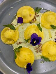 Yellow ravioli plated on a gray dish with creamy herb sauce, toasted pine nuts, fresh mint leaves, olive oil drizzle and bright purple edible pansy flowers — gourmet Italian pasta presentation.