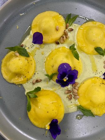 Yellow ravioli plated on a gray dish with creamy herb sauce, toasted pine nuts, fresh mint leaves, olive oil drizzle and bright purple edible pansy flowers — gourmet Italian pasta presentation.