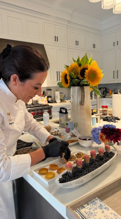 Chef in a white jacket with gloved hands assembling gourmet bite-sized appetizers on a modern home kitchen island, sunflowers in a vase, white cabinets and stainless refrigerator