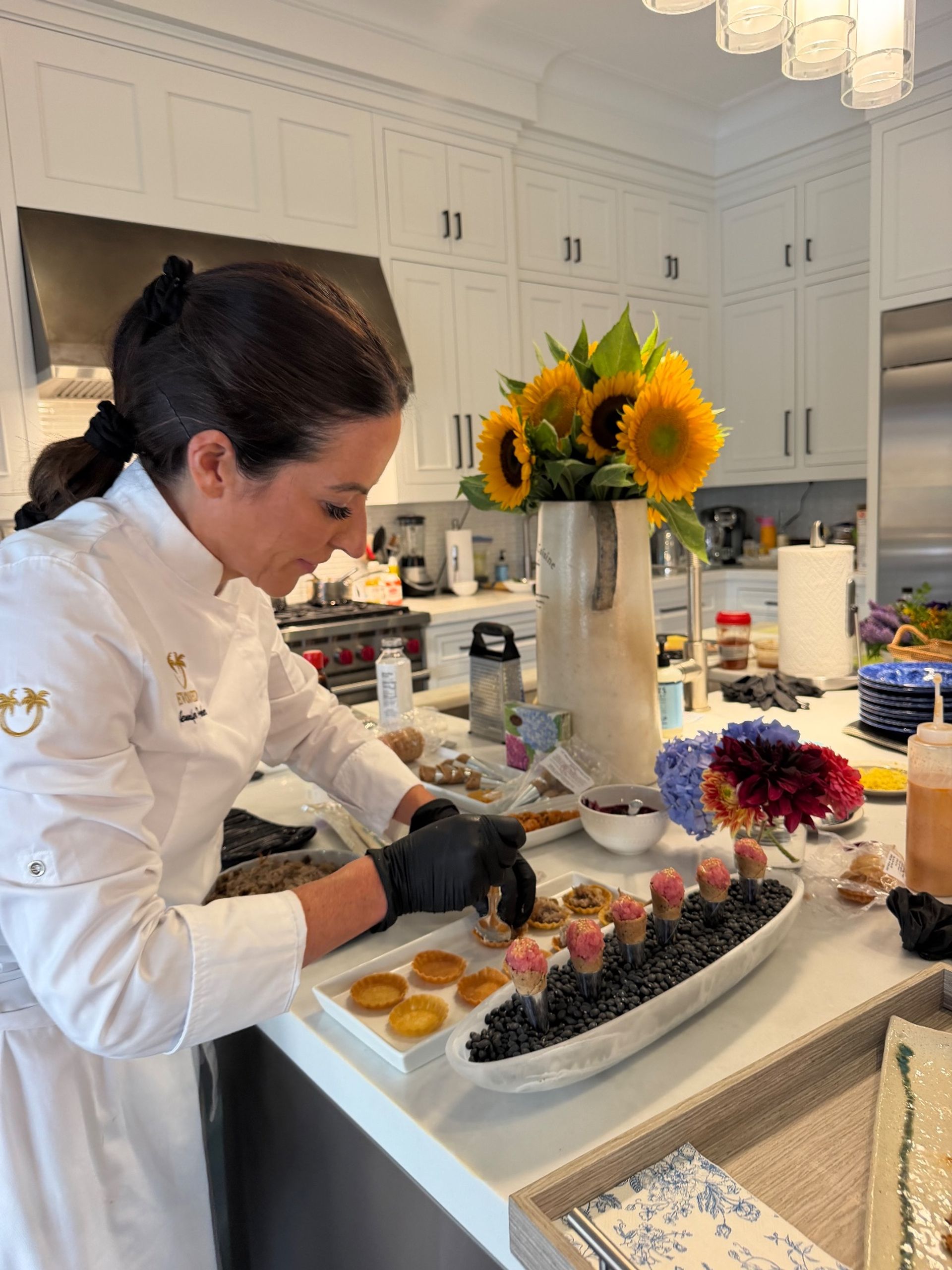 Chef in a white jacket with gloved hands assembling gourmet bite-sized appetizers on a modern home kitchen island, sunflowers in a vase, white cabinets and stainless refrigerator