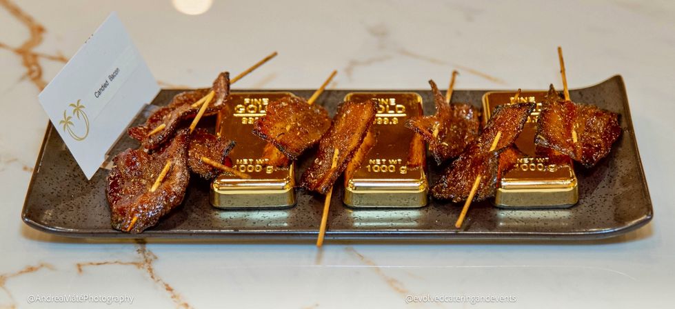 Candied bacon skewers arranged on miniature gold bar props on a black rectangular platter set on a white marble countertop — sweet, sticky luxury appetizer