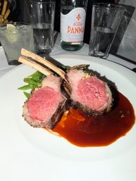 Sliced medium-rare rack of lamb (two bones) on a white plate with glossy red wine jus and green beans, water glasses and a bottle in the background — fine dining dinner.