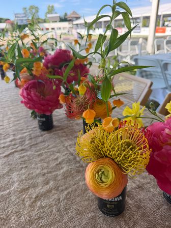 Vibrant floral centerpiece on a linen-covered outdoor patio table featuring pink peonies, yellow pincushion protea, orange ranunculus and small hanging orange lantern blooms in small black vases.