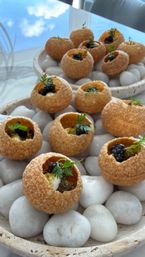 Golden crispy choux puff canapés filled with creamy mousse, caviar and microherbs, arranged on smooth white stones in a shallow ceramic dish — elegant gourmet appetizer for fine dining.