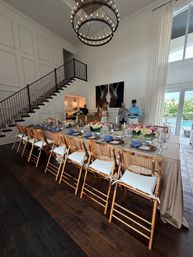 Long banquet table set for dinner in a bright modern dining room — rattan chairs with white cushions, linen table with blue napkins and colorful floral centerpieces, two-tier round chandelier, paneled wall and staircase, glass doors opening to a backyard pool.
