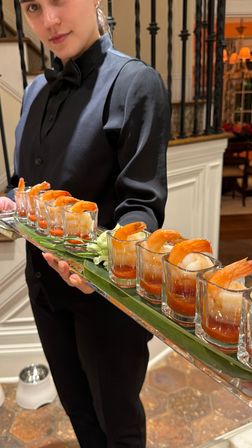 Server in black uniform holding a mirrored tray of shrimp cocktail shooters — row of shot glasses with large shrimp and cocktail sauce, garnished with greens, served at an upscale indoor event near a staircase.