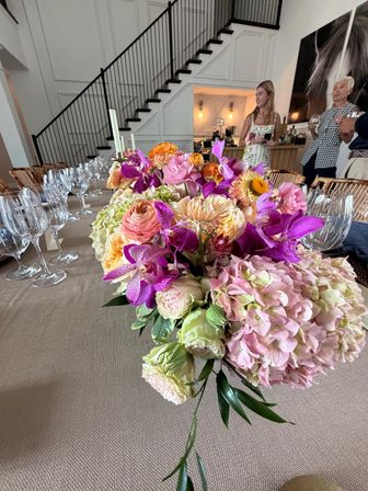 Indoor dining tablescape with a vibrant centerpiece of pink hydrangeas, peach ranunculus and purple orchids, rows of wine glasses and candles, set beneath a modern black staircase with guests in the background.