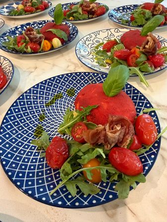 Plated appetizer: red beet dome topped with basil, prosciutto rosette, roasted cherry tomatoes and arugula on blue patterned plates set on a marble countertop
