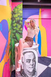 Blonde woman in a sparkly top and brown mini skirt sitting on an oversized rolled $100 prop inside a colorful pop-art installation with pink, yellow and blue mural walls, black-and-white checkerboard floor, and white platform heels
