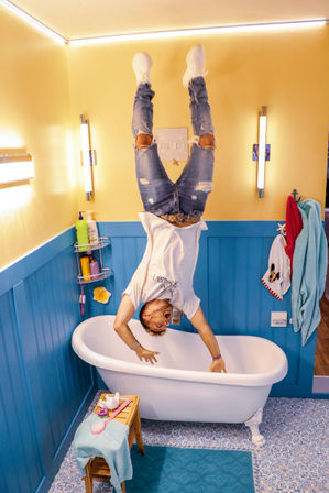 Playful upside-down person in ripped jeans and white sneakers appearing to stand on the ceiling above a white clawfoot bathtub in a bright yellow and blue bathroom with towels and toiletries.