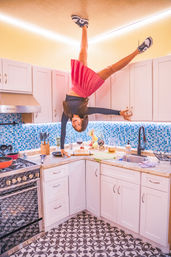 Playful upside-down woman balancing on her hands against white kitchen cabinets in a bright modern kitchen with blue mosaic backsplash, patterned tile floor, stove, sink, and a cake on the countertop.
