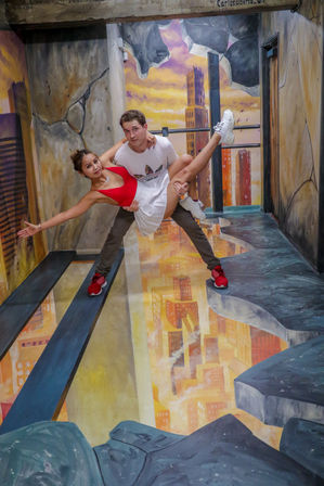 Couple posing in an interactive 3D mural exhibit — man lifts woman in a dance-like pose over a painted crumbling skyscraper and cityscape chasm with reflective floor; woman in red top and white skirt, both wearing sneakers.
