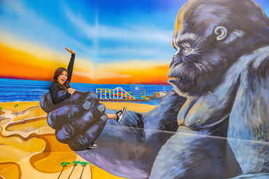 Person posing in a playful 3D mural of a giant gorilla cradling them above a sunlit beach with an amusement pier and Ferris wheel at sunset