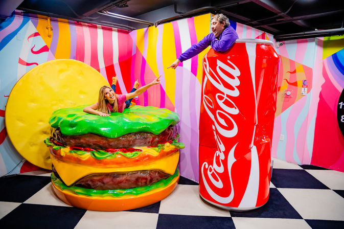 Playful indoor interactive art exhibit: a woman sprawled on a giant cheeseburger prop and a man leaning out of an oversized red soda can, pointing at each other against vibrant striped walls and a black-and-white checkered floor — colorful pop-art photo-op.