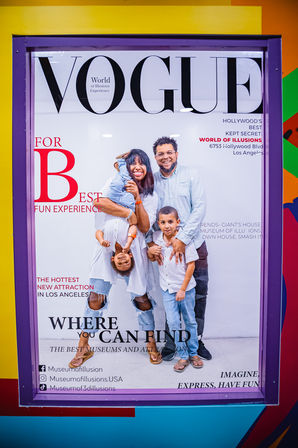 Playful poster of a smiling family striking illusion poses—one parent holding a child upside-down—inside a purple-framed display promoting a Los Angeles museum photo‑op and family attraction.