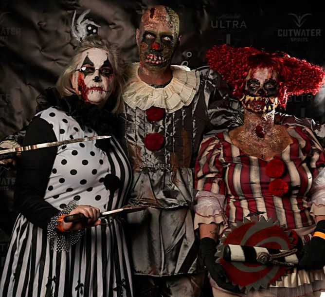 Three people in gruesome, bloody clown Halloween costumes and makeup posing with prop knives and a circular saw in front of a haunted event photo backdrop