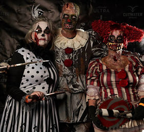 Three people in gruesome, bloody clown Halloween costumes and makeup posing with prop knives and a circular saw in front of a haunted event photo backdrop