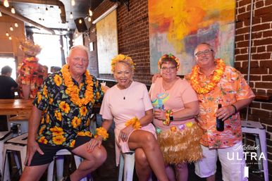 Group of four friends in leis and tropical outfits posing at a luau-themed indoor bar with exposed brick, colorful wall art and hanging lights, one holding a bottle and another a cocktail.