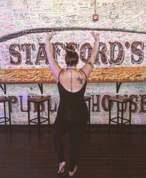 Person with hair in a bun and visible back tattoos, back to camera with arms raised, posing in flip-flops inside a bar/pub interior in front of a colorful, signature-covered brick mural and wooden stools.