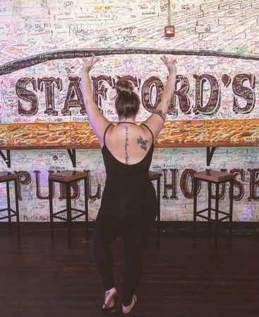 Person with hair in a bun and visible back tattoos, back to camera with arms raised, posing in flip-flops inside a bar/pub interior in front of a colorful, signature-covered brick mural and wooden stools.