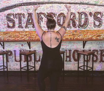 Person with hair in a bun and visible back tattoos, back to camera with arms raised, posing in flip-flops inside a bar/pub interior in front of a colorful, signature-covered brick mural and wooden stools.