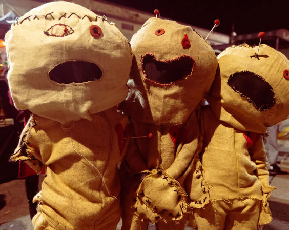 Three burlap voodoo-style dolls with stitched faces, button eyes and pins in their heads, posed together in a nighttime outdoor Halloween display.