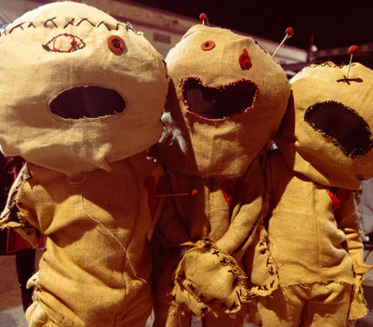 Three burlap voodoo-style dolls with stitched faces, button eyes and pins in their heads, posed together in a nighttime outdoor Halloween display.