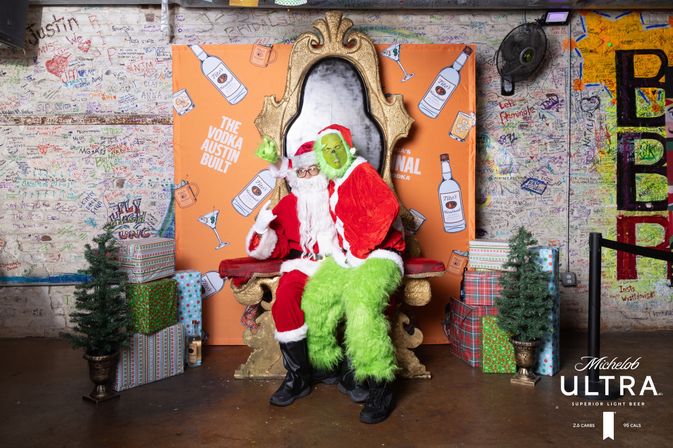 Two costumed characters — Santa and the Grinch — sit on a gold throne in a graffiti-covered indoor Austin holiday photo-op, surrounded by wrapped gifts, small evergreen trees and a festive backdrop.
