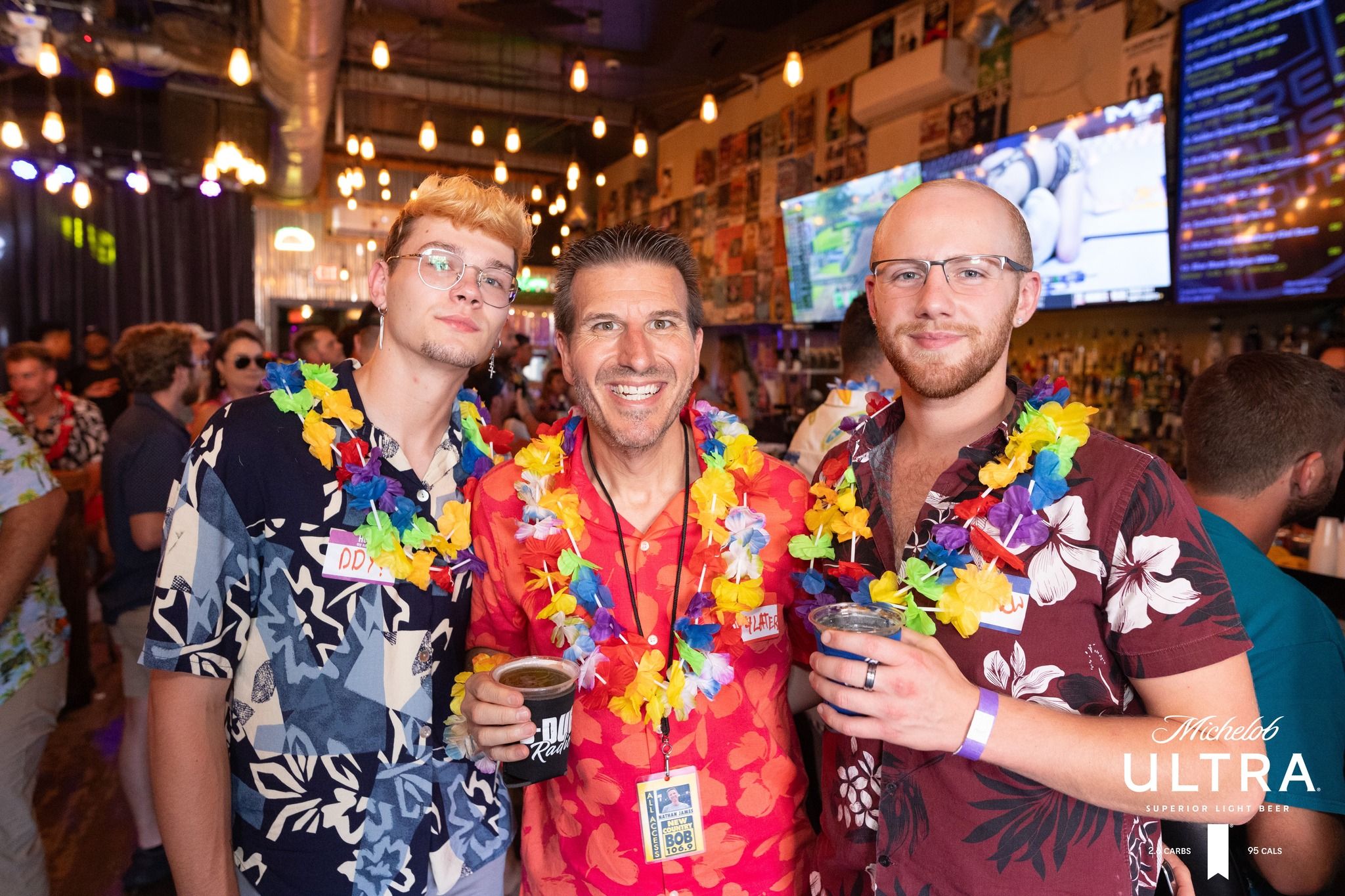Three people in Hawaiian shirts and colorful leis holding drinks and smiling at a lively indoor bar party under string lights