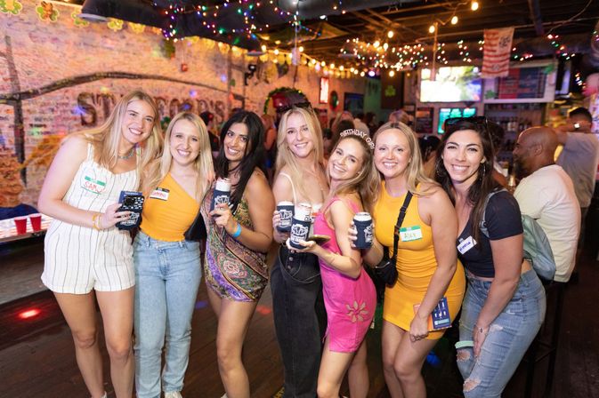 Eight friends smiling and posing with canned drinks in koozies inside a neon-lit bar with string lights and a graffiti-covered brick wall — lively nightlife scene and birthday celebration.