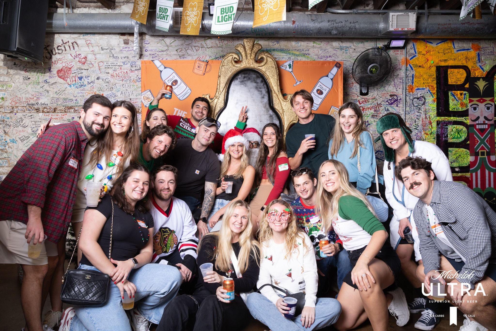 Large group of friends posing at a festive bar photo wall — colorful graffiti background and ornate gold throne prop; holiday hats, novelty glasses and drinks in hand at a lively holiday party.