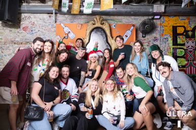 Large group of friends posing at a festive bar photo wall — colorful graffiti background and ornate gold throne prop; holiday hats, novelty glasses and drinks in hand at a lively holiday party.